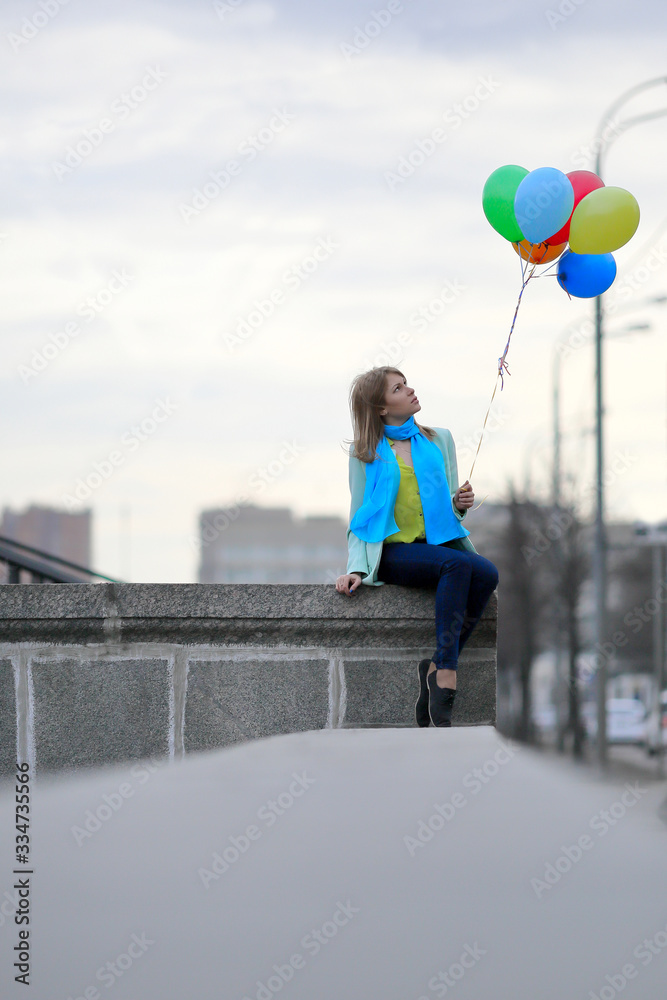 Town. Early spring. Gray city landscape. A girl with a blue scarf in bright clothes walks with balloons.
