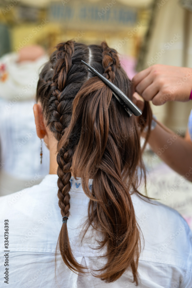 Fototapeta premium Super styling. Rear view closeup of a hairdresser braiding her clients hair in trendy weave plait while open-air festival