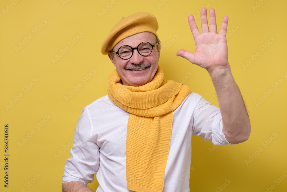 Naklejka premium Senior man in glasses and yellow hat and scarf saying hello, waving a hand.