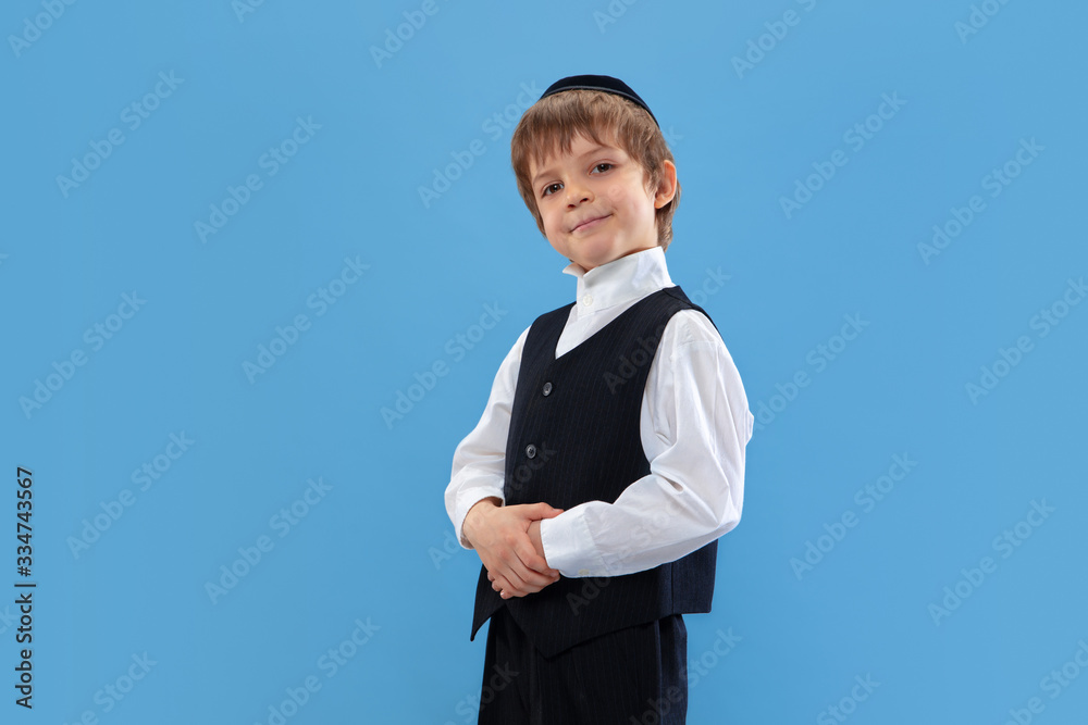 Posing confident, cute. Portrait of a young orthodox jewish boy isolated on blue studio background. Purim, business, festival, holiday, celebration Pesach or Passover, judaism, religion concept.