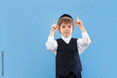 Pointing. Portrait of a young orthodox jewish boy isolated on blue studio background. Purim, business, festival, holiday, childhood, celebration Pesach or Passover, judaism, religion concept.