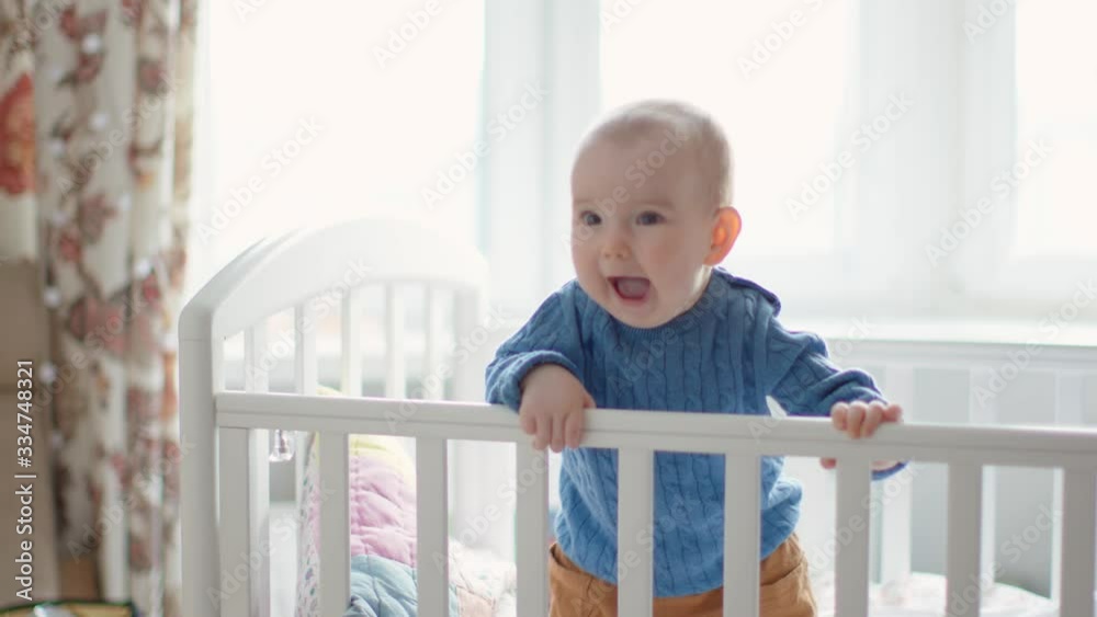 Cheerful Infant Baby Learns to Stand on his Feet in a Crib. Sunny Home Interior. Cheerful Infant Baby Learns to Stand on his Feet in a Crib. Sunny Home Interior.