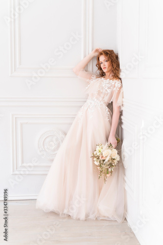 Beautiful natural redhead girl bride, with nude makeup, wearing a white dress, holds a wedding bouquet in her hands, standing an the wall in a light interior.