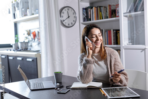 Giovane donna pratica lo smart working da casa, e parla al telefono sorridendo , sullo sfondo la cucina photo