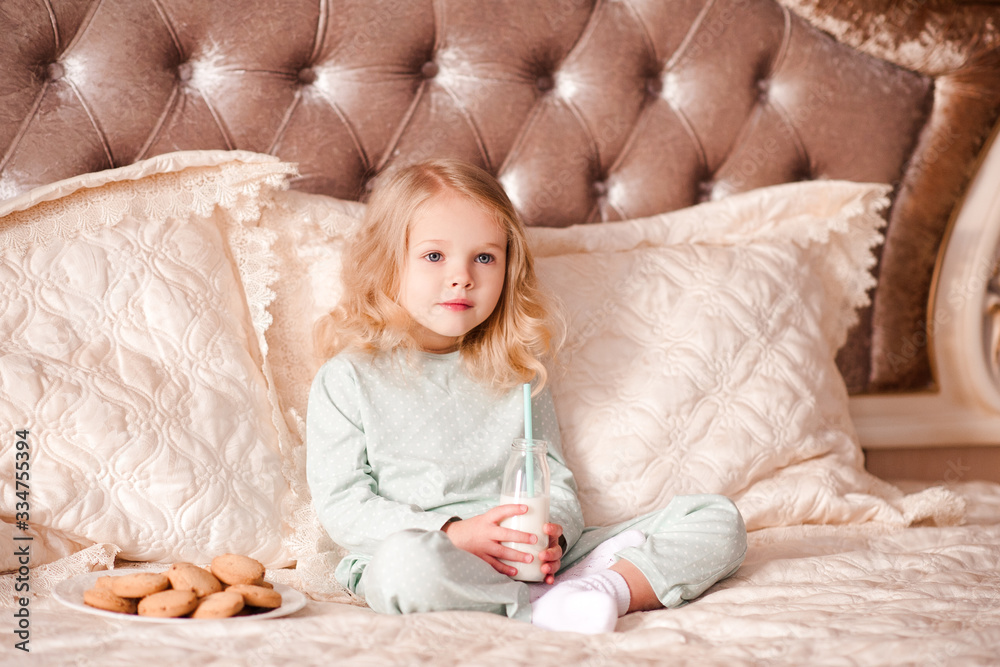 Fototapeta premium Cute baby 3-4 year old drinking milk and eating cookies in bed closeup. Breakfast time. Good morning. Childhood.