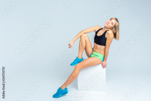 Portrait of beautiful athletic caucasian woman in sport bra and shorts sitting on a white cube on a white studio backgroung. Copy space