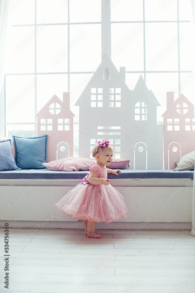 Fototapeta premium Portrait of a happy little blonde girl in a pink dress sitting on the windowsill