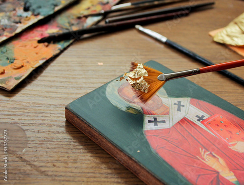 Hand with brush of gold leaf. Artist applies gold leaf to the icon. Reproduction of the icon of St. Nicholas. The process of the artist's work. Brushes and paints on the background. photo