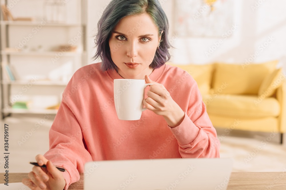 Selective focus of of freelancer with colorful hair looking at camera and holding cup of tea with pen at table in living room