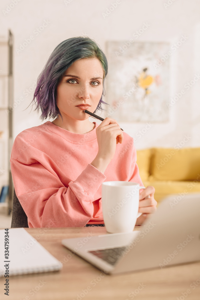 Selective focus of freelancer with colorful hair looking at camera and holding pen at table Selective focus of freelancer with colorful hair looking at camera and holding pen at table