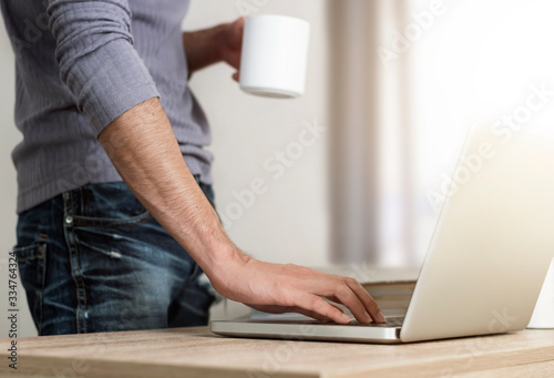 Close up low angle view of a man standing at the a desk surfing on internet and drinking coffee in morning. photo