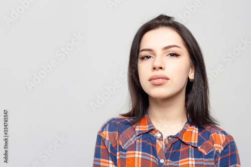 Portrait young beautiful woman not smiling in plaid shirt isolated on white-gray background. Girl with minimal natural make-up on her face. The photo is not retouched with natural defects on the skin. © simikov