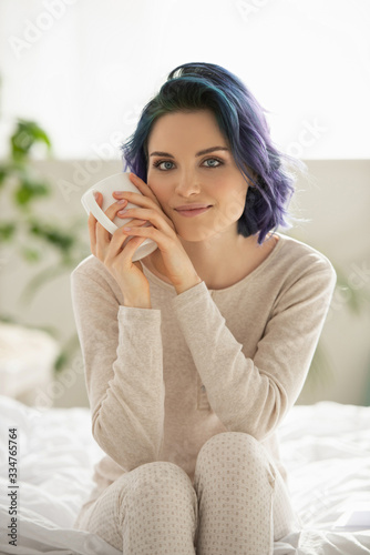 Beautiful girl with colorful hair holding cup of tea, looking at camera and smiling on bed in bedroom