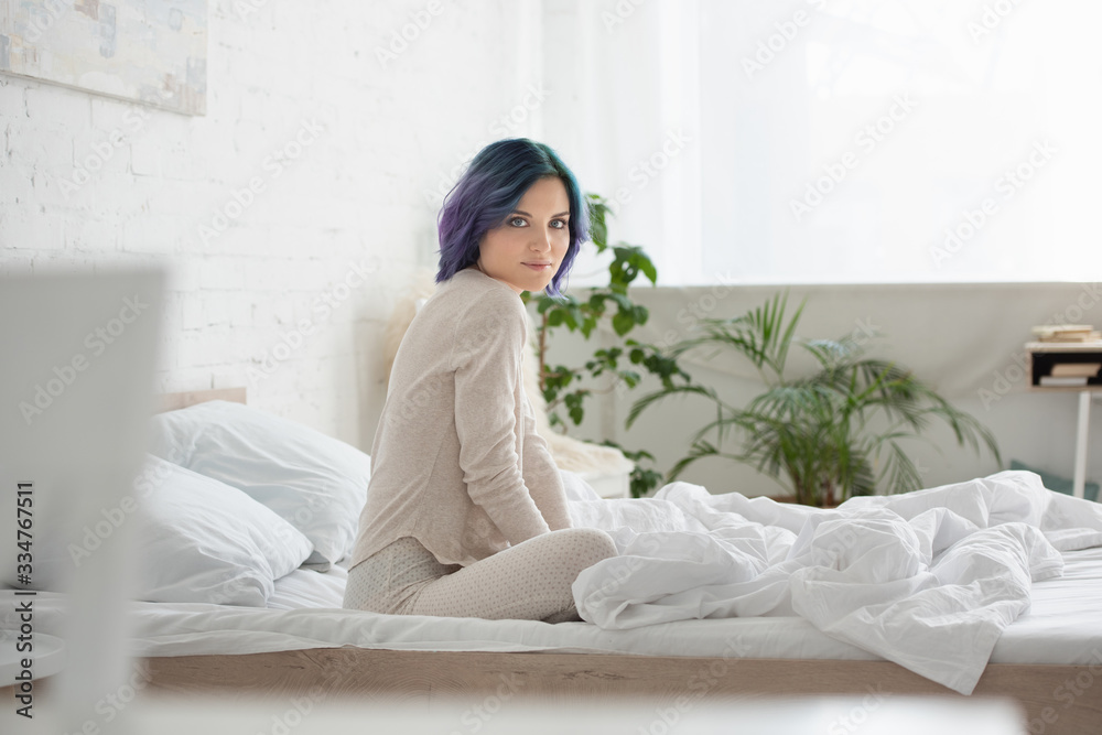 Selective focus of woman with colorful hair looking at camera and sitting on bed in bedroom Selective focus of woman with colorful hair looking at camera and sitting on bed in bedroom