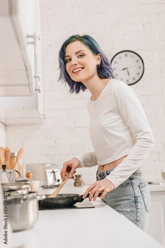 Low angle view of girl with colorful hair preparing food with spatula on frying pan, smiling and looking at camera in kitchen Low angle view of girl with colorful hair preparing food with spatula on frying pan, smiling and looking at camera in kitchen