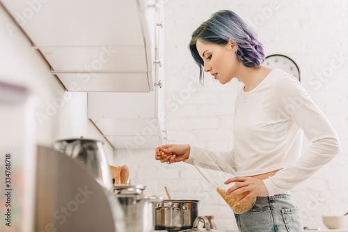 Selective focus of girl with colorful hair putting pasta in pan near kitchen stove