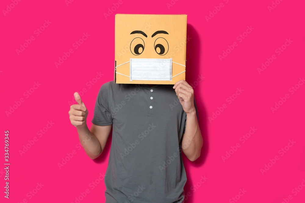 Man wearing cardboard box with mask to protect from COVID 19 virus showing thumbs up sign against pink background
