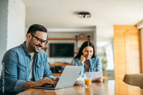 Portrait of a handsome smiling man using laptop, woman in the background reading letter.