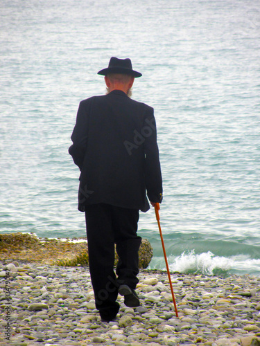 Slika na platnu An elderly man in a black suit with a cane walks along the sea pebble beach