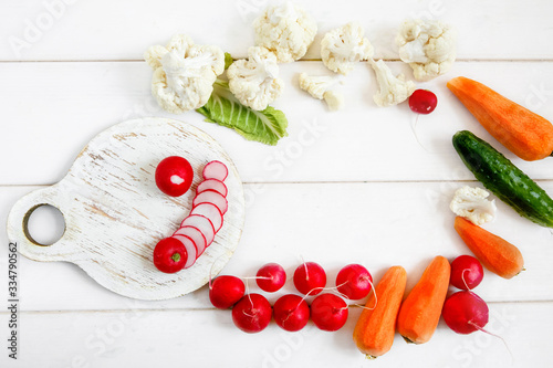Fresh vegetables on a white wooden table - cauliflower, radishes, cucumbers and carrots. On a round cutting Board sliced radishes