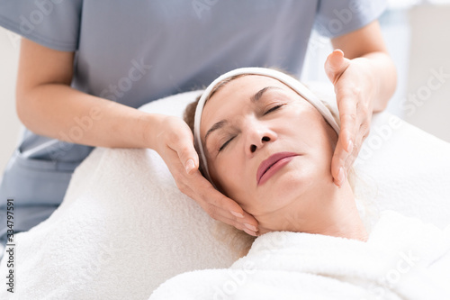Close-up of beautician massaging face of mature female client relaxing at procedure