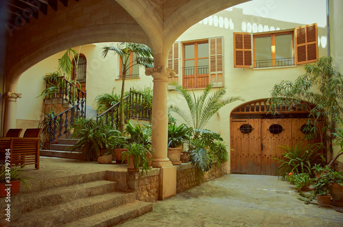 Inner courtyard with lot of plants. There is a porch with a wooden table and chairs. There are two wooden windows  a wooden door and stairs to go to the main door. Mallorca  Spain 08 17