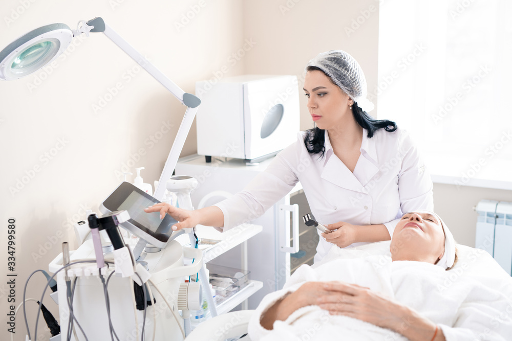 Cosmetologist in surgical cap choosing power for face peeling on machine while preparing to do cosmetic procedure in cosmetology room Cosmetologist in surgical cap choosing power for face peeling on machine while preparing to do cosmetic procedure in cosmetology room