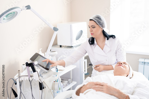 Cosmetologist in surgical cap choosing power for face peeling on machine while preparing to do cosmetic procedure in cosmetology room