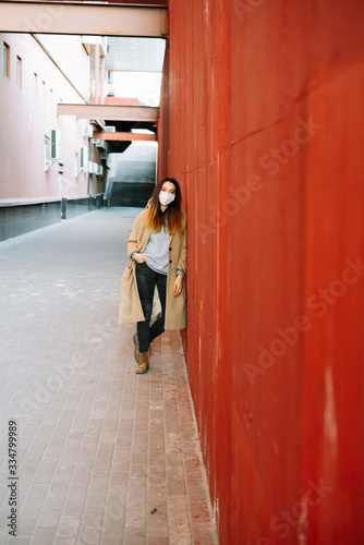 a young girl in a brown coat with a medical mask on her face,fighting against the corona 19 virus infection on the street, near the red wall © Дарья Фомина
