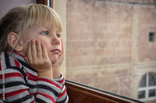 The white-haired blue-eyed girl looks out the window. Quarantined child. The child is sad at the window. photo