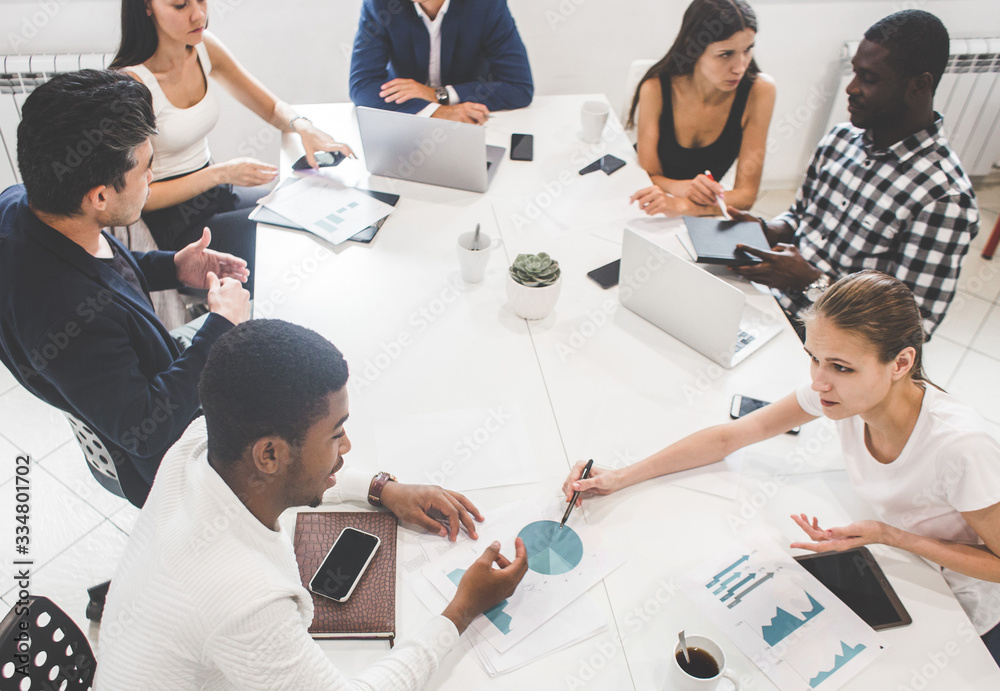 © xartproduction - A team of young office workers, businessmen with laptop working at the table, communicating together in an office. Corporate businessteam and manager in a meeting. coworking.