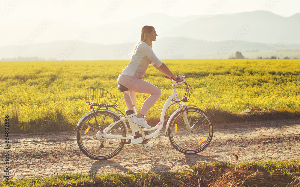 Young woman rides electric bicycle on dusty country road, view from side, sun backlight field with yellow flowers background