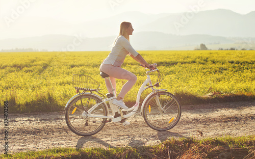 Young woman rides electric bicycle on dusty country road, view from side, sun backlight field with yellow flowers background