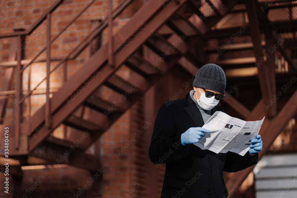 Coronavirus precautions. Male model avoids crowded places, fights against new virus, reads attentively article about covid-19 symptoms, holds newspaper, wears protective face mask and medical gloves Coronavirus precautions. Male model avoids crowded places, fights against new virus, reads attentively article about covid-19 symptoms, holds newspaper, wears protective face mask and medical gloves
