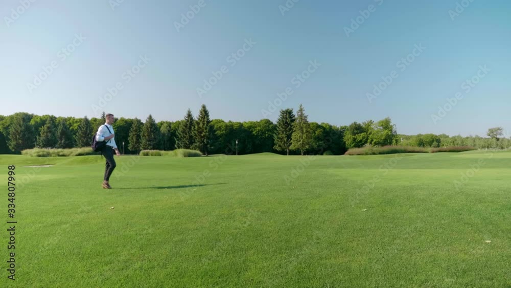 Young businessman with a backpack on his shoulder walks in the park. Man in a classic white shirt and trousers walks on green grass. 4K footage Young businessman with a backpack on his shoulder walks in the park. Man in a classic white shirt and trousers walks on green grass. 4K footage