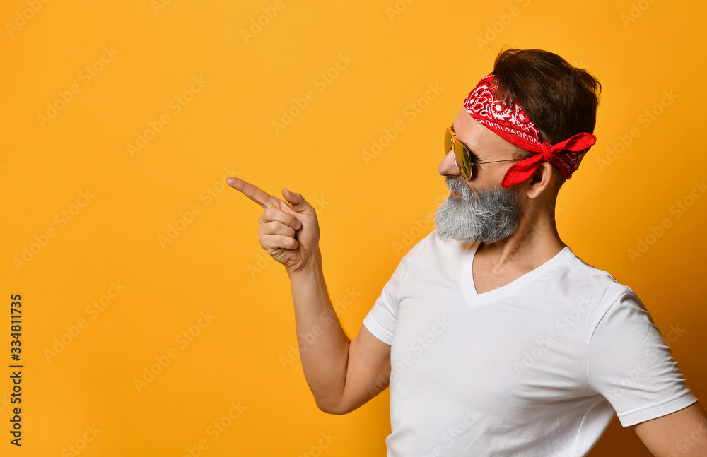 Mature man in sunglasses, red bandana, white t-shirt. Smiling, pointing at something by forefinger, posing on orange background