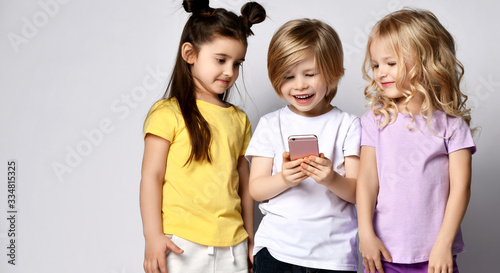 Two little baby girls and boy in stylish casual clothing standing, looking at smartphone screen and smiling