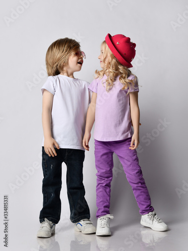 Little positive blond baby girl and boy in stylish casual clothing and accessories standing and looking at each other photo