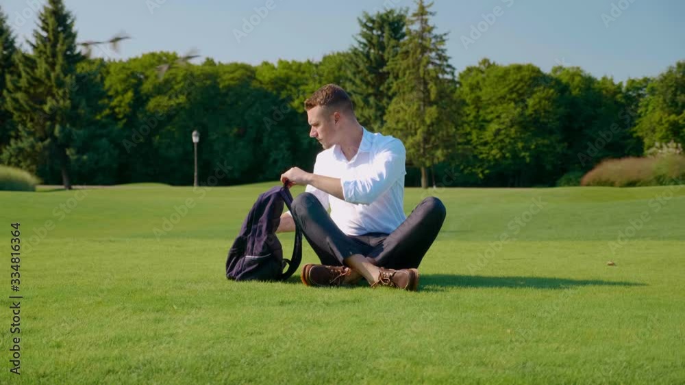 Businessman takes out his vegetarian lunch with eco-packaging from a backpack. A freelancer in a classic white shirt and trousers sits in a park on green grass and takes out a paper package with lunch Businessman takes out his vegetarian lunch with eco-packaging from a backpack. A freelancer in a classic white shirt and trousers sits in a park on green grass and takes out a paper package with lunch