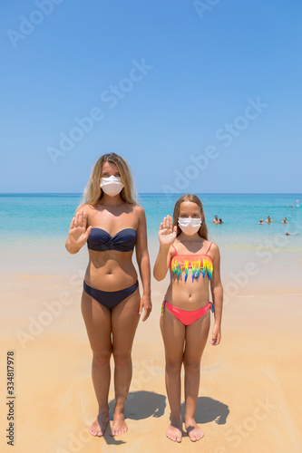 A woman and a child in a protective surgical mask on their face are standing on the beach in swimsuits. Chinese coronavirus disease COVID-19 is dangerous