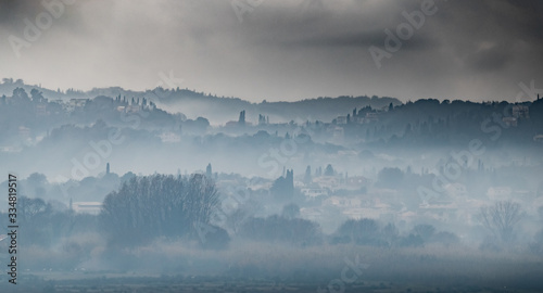 Fog over the city on a slope in cloudy weather, cypresses stick out, Corfu, Greece