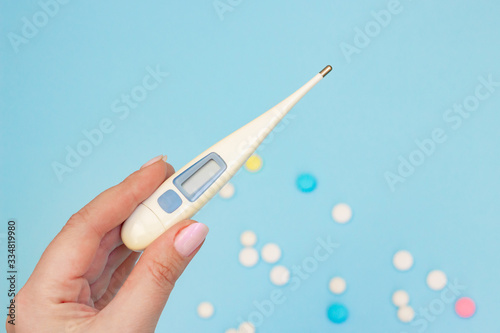Electronic thermometer in hand on a blue background with scattered pills. Elevated body temperature as a symptom of coronavirus infection