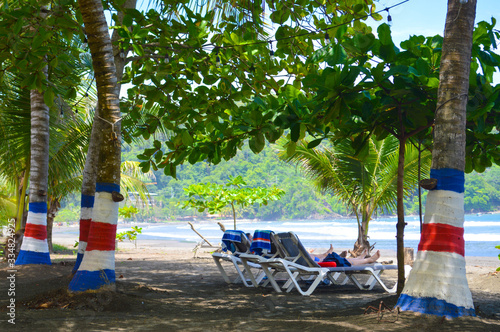 At Jaco beach, Costa Rica. With coloured painted palm trees with the flag of Costa Rica. Tourists relaxing under the shade of two palm trees and an 