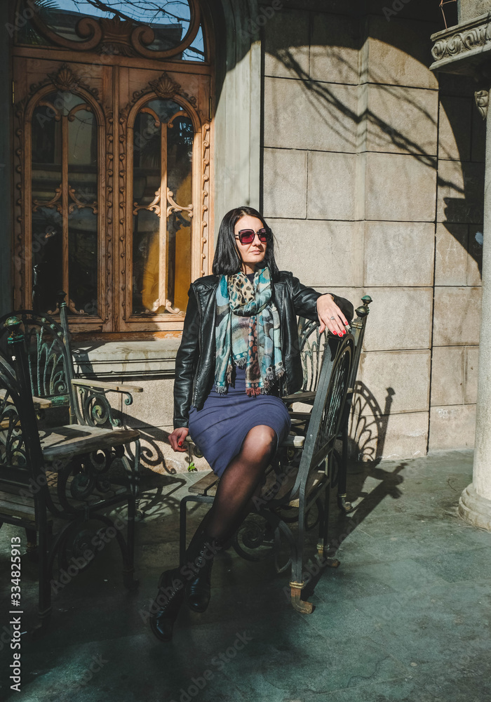 Obraz premium Fashion, brunette woman in a dress and leather black jacket sitting on old iron chair outdoors. Girl model posingl in Batumi, Georgia. Old city at background.