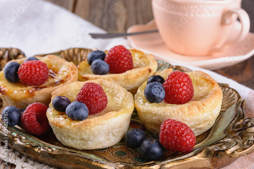 Small puff pastry tartlets stuffed with creme brulee and fresh blueberries and raspberries on a vintage tray next to cup of tea or coffee