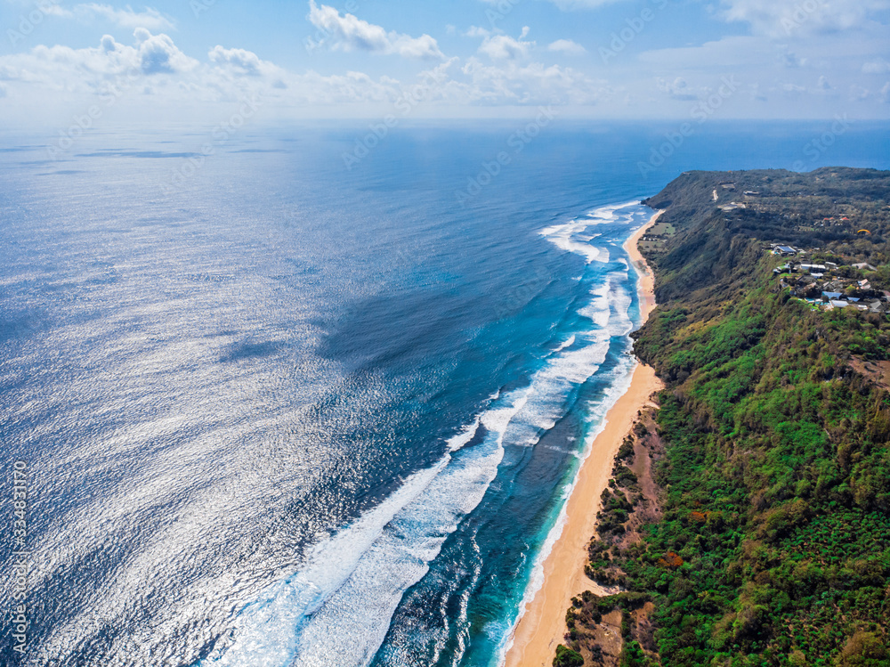Aerial photo of a wild beach in Bali. The southern part of the island of Bali is the Bukit Peninsula. A long strip of beach and a steep mountain slope covered with greenery. Powerful ocean waves roll 