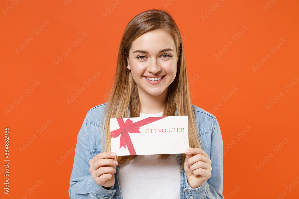 Smiling young woman girl in casual denim clothes posing isolated on bright orange wall background studio portrait. People sincere emotions lifestyle concept. Mock up copy space. Hold gift certificate. Smiling young woman girl in casual denim clothes posing isolated on bright orange wall background studio portrait. People sincere emotions lifestyle concept. Mock up copy space. Hold gift certificate.