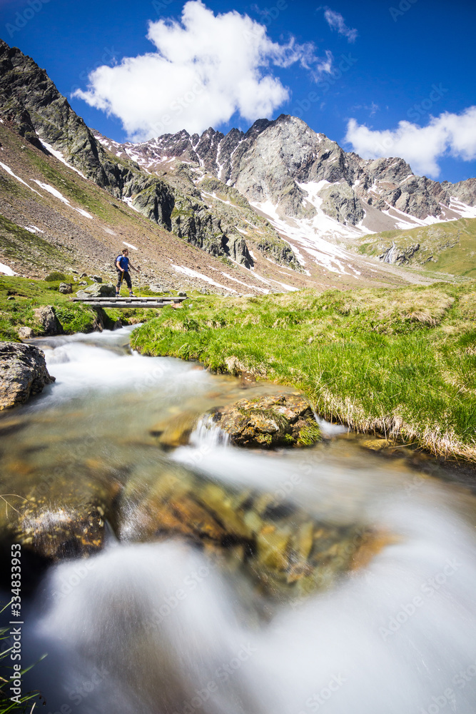 Hiker in the mountains crosses the bridge over the stream in summer, Stelvio Park, Lombardy, Italy