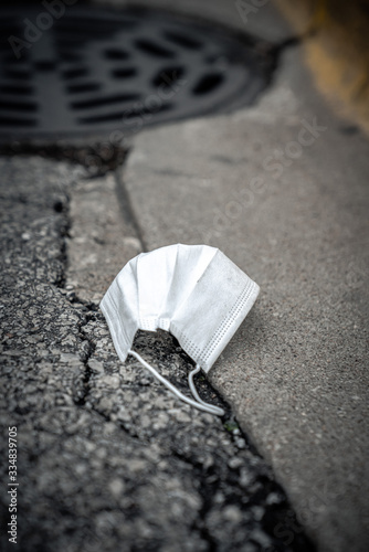 A close up of a dirty used white medical mask or surgical respiratory face mask discarded and laying as trash on asphalt street next to a concrete curb and storm sewer manhole in the city.