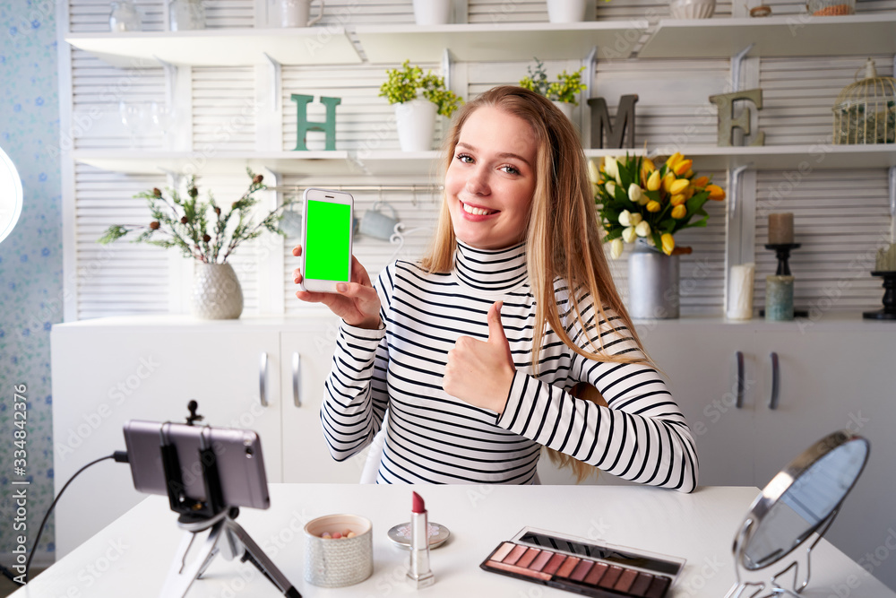 Beauty blogger woman filming advertising, holding green screen smartphone application. Make-up influencer blonde girl live streaming cosmetics product review in studio asking for likes, subscription Beauty blogger woman filming advertising, holding green screen smartphone application. Make-up influencer blonde girl live streaming cosmetics product review in studio asking for likes, subscription